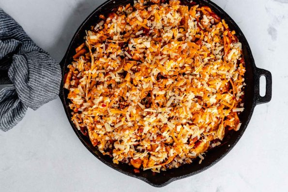 Black bean & butternut squash enchiladas skillet topped with grated cheese. The skillet sits atop a light blue surface. The handle of the skillet is wrapped with a black & white striped linen napkin.