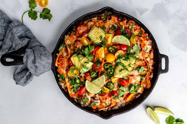 Vegetarian skillet enchiladas, shown in a large black skillet, topped with melted cheese, cherry tomatoes, sliced avocado, roughly chopped cilantro, thinly sliced jalapeno pepper. The skillet sits atop a light blue surface, with its handle wrapped in a black & white linen napkin. Next to the skillet, on the surface, are cilantro leaves & lime wedges for serving.