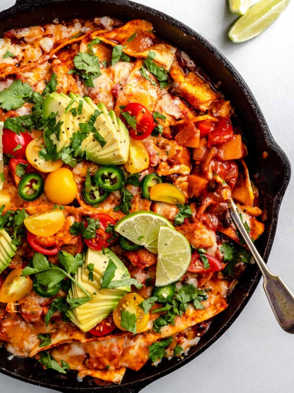 Close up of vegetarian skillet enchiladas, shown in a large black skillet, topped with melted cheese, cherry tomatoes, sliced avocado, roughly chopped cilantro, thinly sliced jalapeno pepper. The skillet sits atop a light blue surface, with its handle wrapped in a black & white linen napkin. Next to the skillet, on the surface, are cilantro leaves & lime wedges for serving.