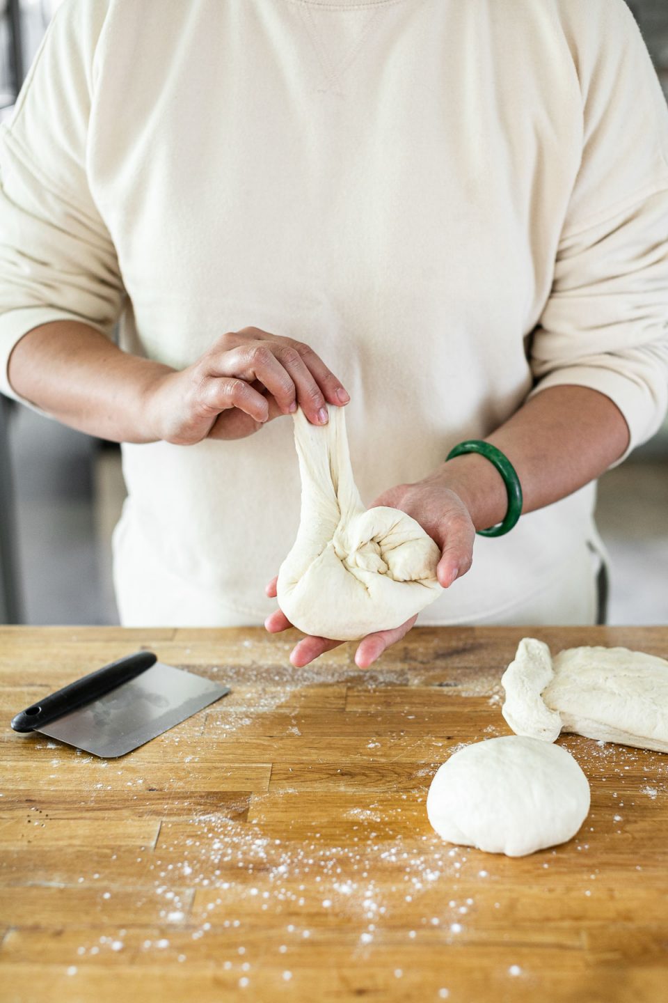 How to shape pizza dough balls: Jess, shown in a white sweatshirt, stands behind a butcher block counter dusted in flour. Jess is folding in edges of the pizza dough, forming a ball.
