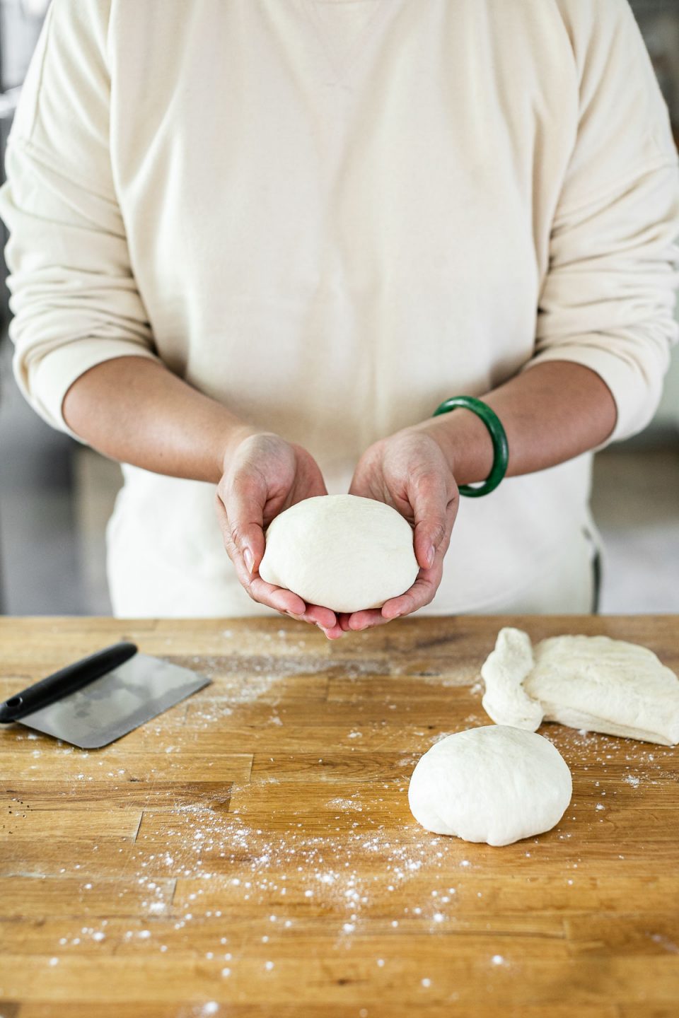 How to shape pizza dough balls: Jess, shown in a white sweatshirt, stands behind a butcher block counter dusted in flour. Jess holds up a formed pizza dough ball in her hands.