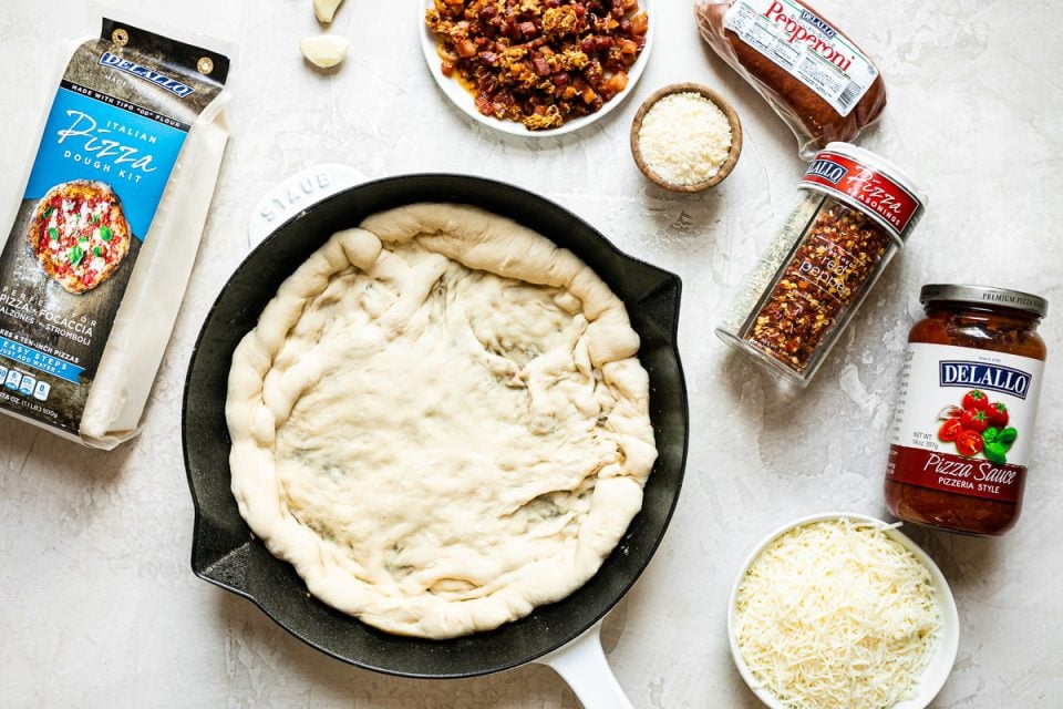 Cast iron skillet pizza ingredients arranged on a white surface: DeLallo Pizza Dough kit, dough formed in a 10-inch white Staub cast iron skillet, garlic cloves, pancetta, parmesan cheese, DeLallo pepperoni, DeLallo pizza seasonings, DeLallo pizza sauce, & shredded Italian cheese.