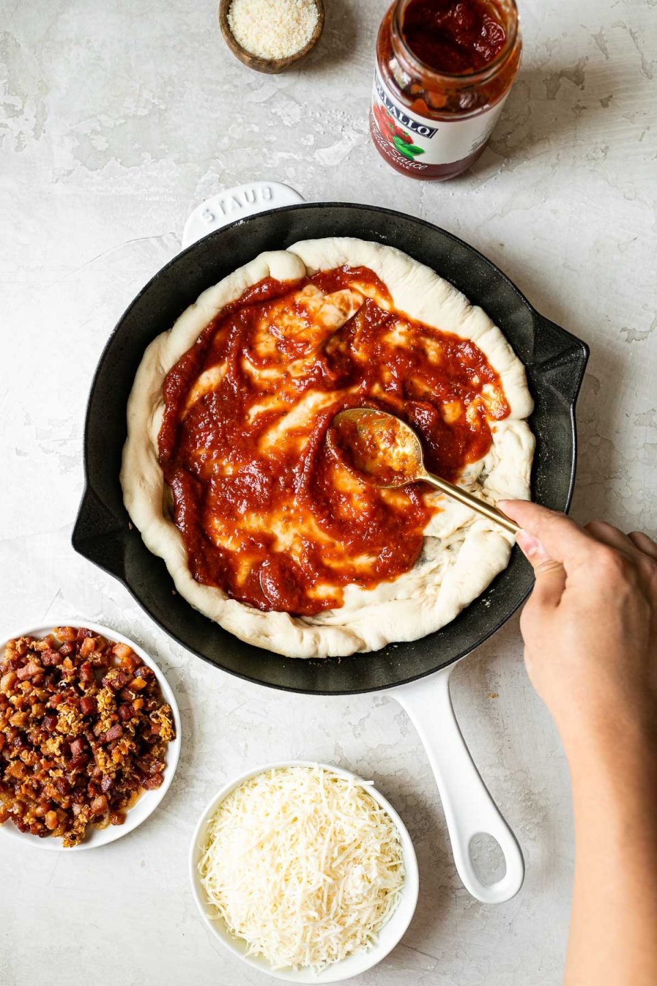 A woman's hand reaching into the frame with a gold spoon, spreading DeLallo pizza sauce over pizza crust formed in a white Staub cast iron skillet. The skillet sits on a white surface, surrounded by parmesan cheese, a jar of DeLallo pizza sauce, fried pancetta & garlic, & shredded Italian cheese.