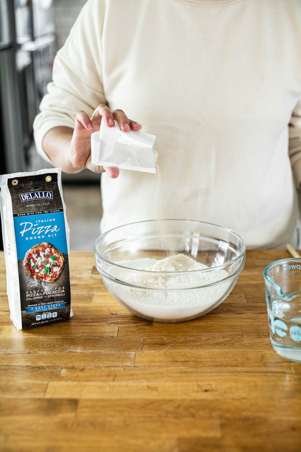 Mixing DeLallo Pizza Dough Kit: Jess, shown in a white sweatshirt, stands behind a butcher block counter, yeast packet into a mixing bowl with flour in it. Sitting next to the bowl on the counter are a wooden spoon & a package of DeLallo Pizza Dough Kit.
