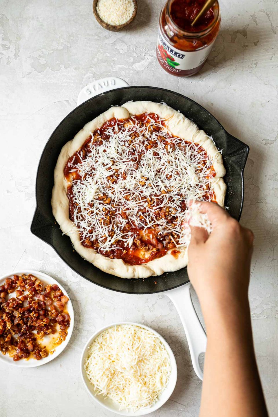 A woman's hand reaching into the frame sprinkling shredded cheese over pizza crust formed in a white Staub cast iron skillet. The skillet sits on a white surface, surrounded by parmesan cheese, a jar of DeLallo pizza sauce, fried pancetta & garlic, & shredded Italian cheese.