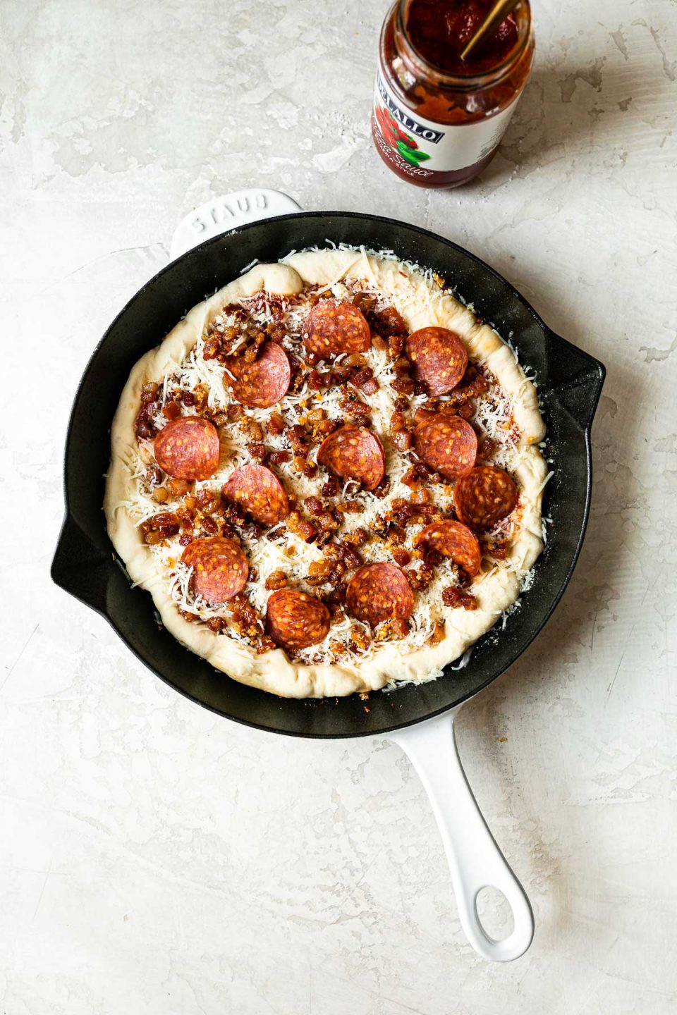 Pizza Amatriciana formed in white Staub cast iron skillet, topped with pepperoni. The skillet sits on a white surface, surrounded by parmesan cheese, a jar of DeLallo pizza sauce.