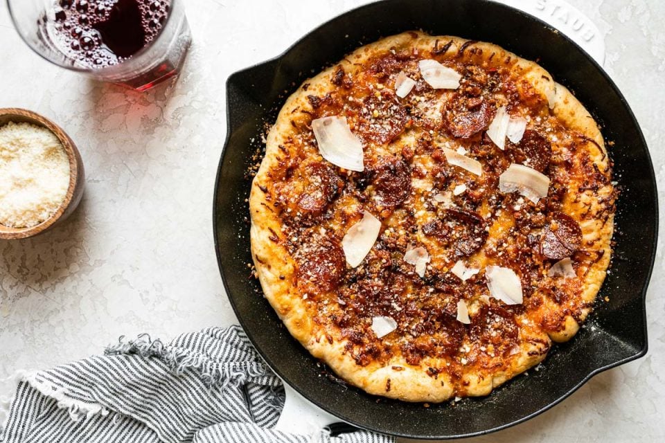 Baked Pizza Amatriciana in white Staub cast iron skillet. The skillet sits on a white surface, surrounded by a striped linen napkin, a glass of red wine & a small wooden bowl filled with grated parmesan cheese.