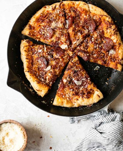 Baked Pizza Amatriciana in white Staub cast iron skillet. The pizza is cut into 6 slices. The skillet sits on a white surface, surrounded by a striped linen napkin, a glass of red wine & a small wooden bowl of grated parmesan cheese.
