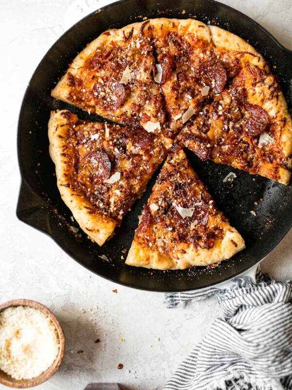 Baked Pizza Amatriciana in white Staub cast iron skillet. The pizza is cut into 6 slices. The skillet sits on a white surface, surrounded by a striped linen napkin, a glass of red wine & a small wooden bowl of grated parmesan cheese.