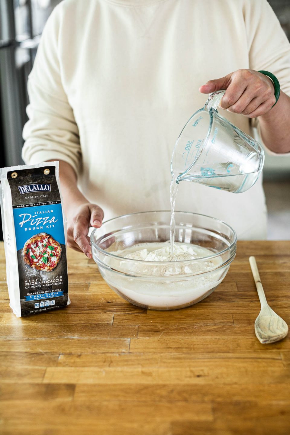Mixing DeLallo Pizza Dough Kit: Jess, shown in a white sweatshirt, stands behind a butcher block counter, pouring water into a mixing bowl with flour in it. Sitting next to the bowl on the counter are a wooden spoon & a package of DeLallo Pizza Dough Kit.