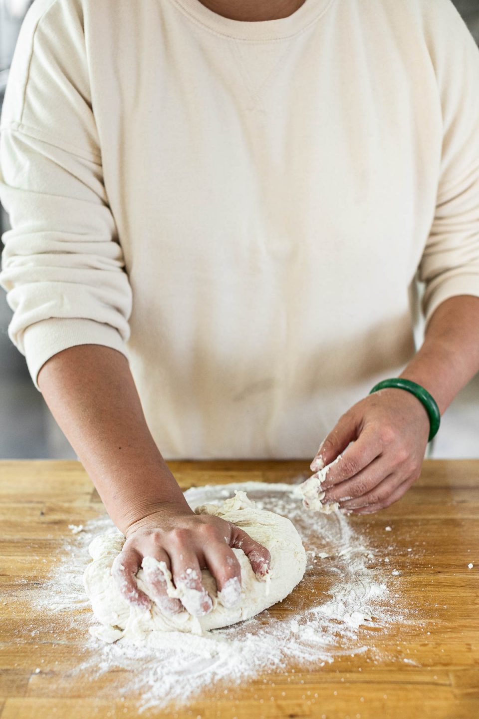 Mixing DeLallo Pizza Dough Kit: Jess, shown in a white sweatshirt, stands behind a butcher block counter, kneading the pizza dough with her hands.