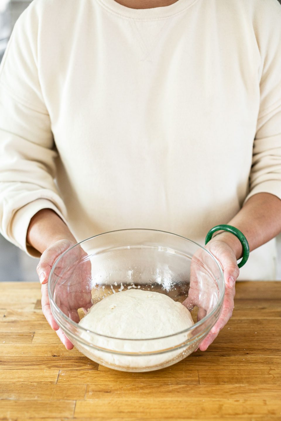 Mixing DeLallo Pizza Dough Kit: Jess, shown in a white sweatshirt, stands behind a butcher block counter holding a large glass mixing bowl with the kneaded dough inside.