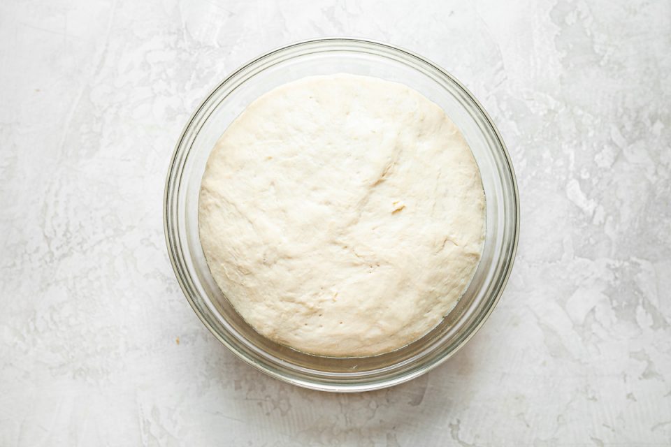 Risen DeLallo Pizza Dough shown in a clear glass mixing bowl atop a white surface.