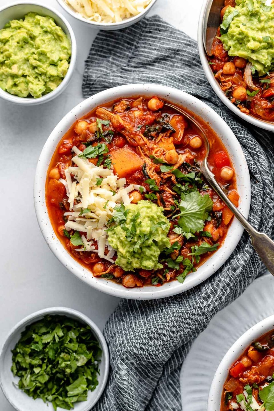 A white bowl filled with Chickpea Chipotle Turkey Chili sits on top of a light blue background. Small bowls of toppings surround the bowl including guacamole, cheese, and cilantro and a blue and white striped napkin is tucked alongside the bowl. A spoon is nestled into the bowl of chili and the chili is topped with cheese, cilantro, and guacamole. 