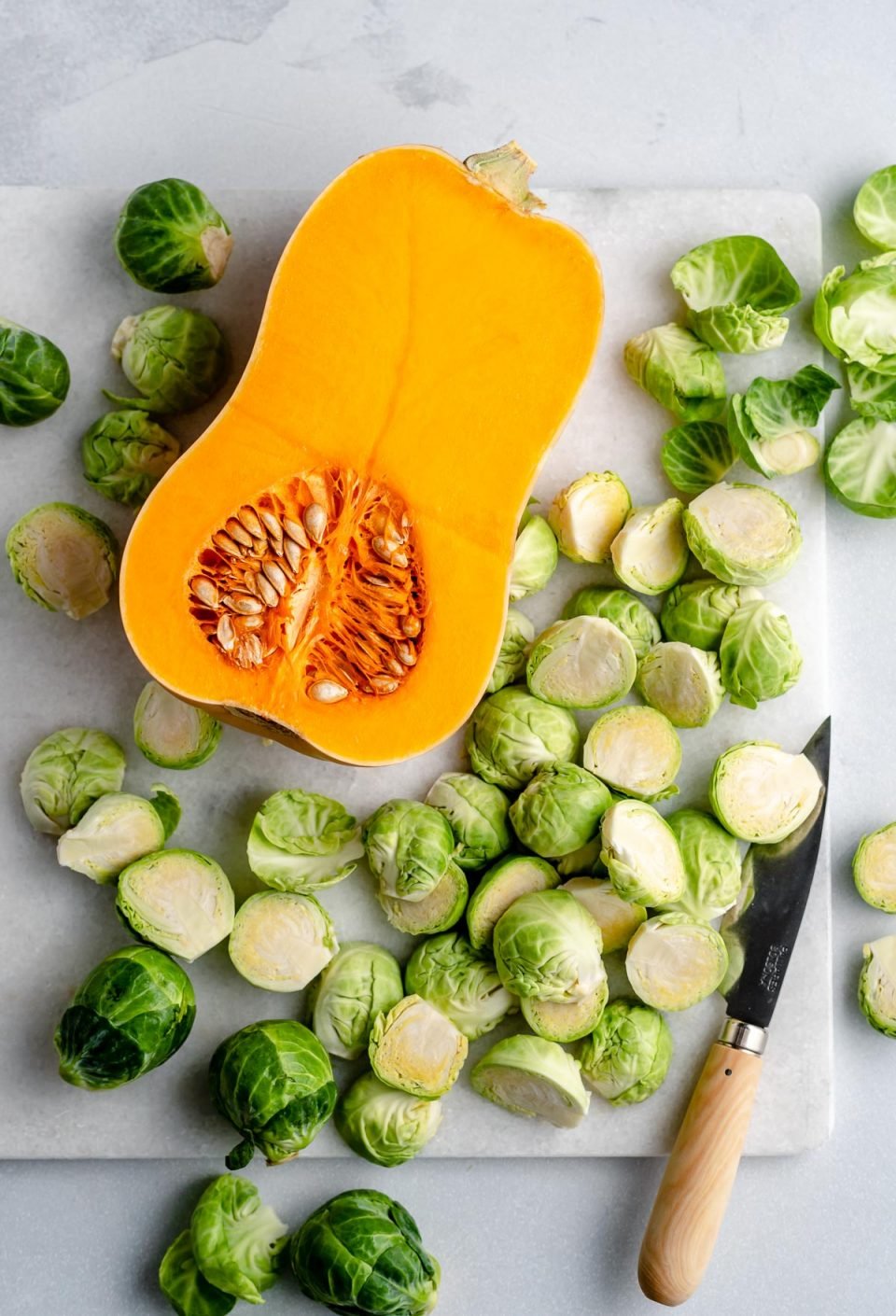Chicken sausage sheet pan prep: a peeled & halved butternut squash with trimmed & halved Brussels sprouts on a white cutting board with a small paring knife.