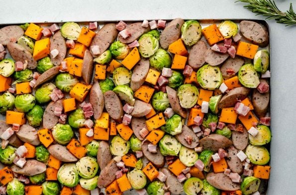 Sheet pan chicken sausage & veggies before roasting - sliced chicken sausage, diced butternut squash, halved brussels sprouts, & diced pancetta on a large sheet pan, topped with finely chopped fresh herbs.