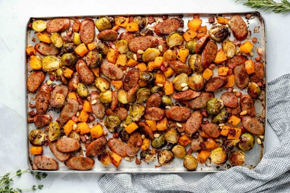 Sheet pan chicken sausage & vegetables. Thinly sliced chicken sausage, diced butternut squash, & halved brussels sprouts are browned & roasty on a large sheet pan. The chicken sausage sheet pan sits atop a light blue surface with a striped linen napkin.