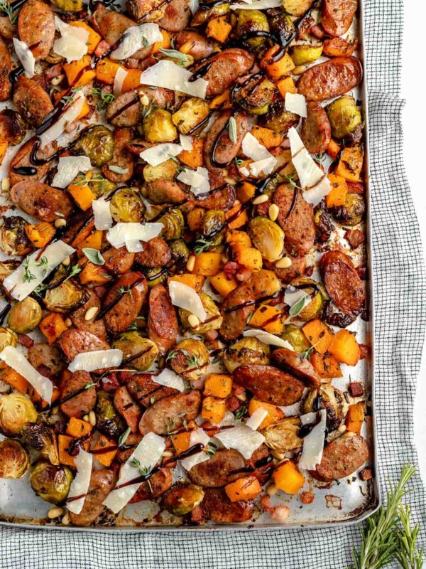 Sheet pan chicken sausage & vegetables. Thinly sliced chicken sausage, diced butternut squash, & halved brussels sprouts are browned & roasty on a large sheet pan, & topped with grated parmesan & a drizzle of balsamic glaze. The chicken sausage sheet pan sits atop a light blue surface with a striped linen napkin.