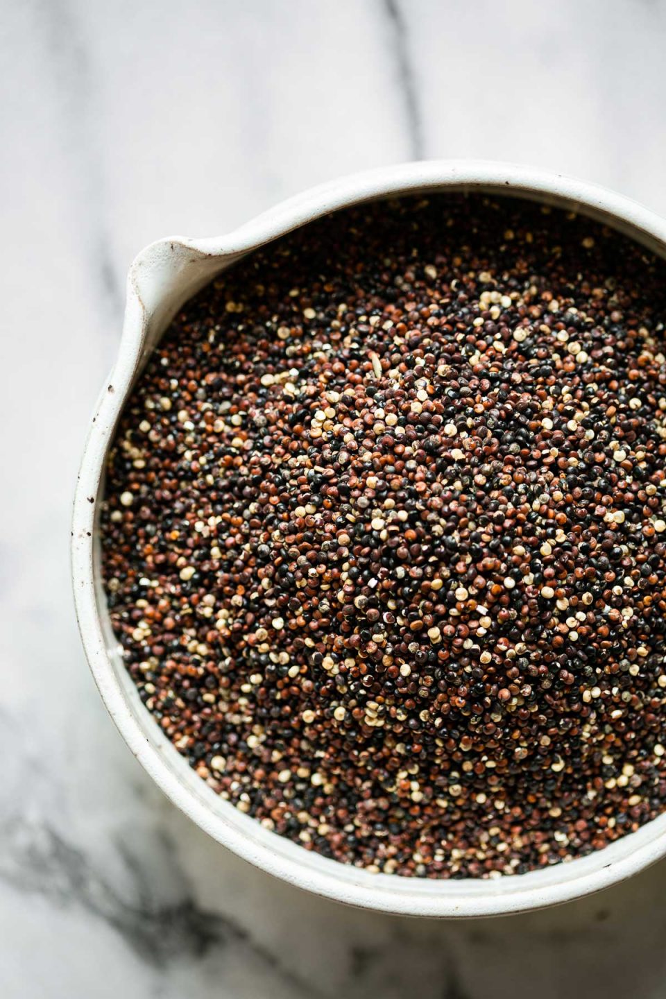 A close up of a small white ceramic bowl filled black quinoa. The bowl sits on top of a white and gray marble surface.