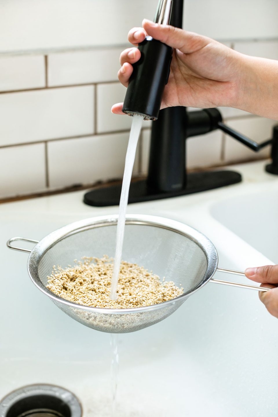 A woman's hand rinses a cup of white quinoa in a fine mesh sieve. Jess is holding the sieve over the sink as the sink sprayer rinses the quinoa with water that is draining through the sieve into the sink.