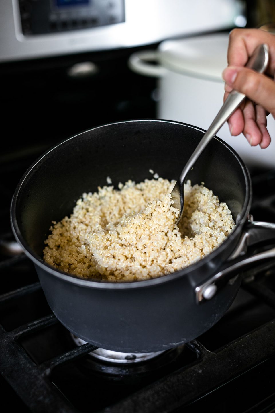 A sauce pan filled with cooked white quinoa sits on top of a gas stovetop. A woman's hand is holding a fork & fluffing the perfectly cooked quinoa prior to serving or storing.