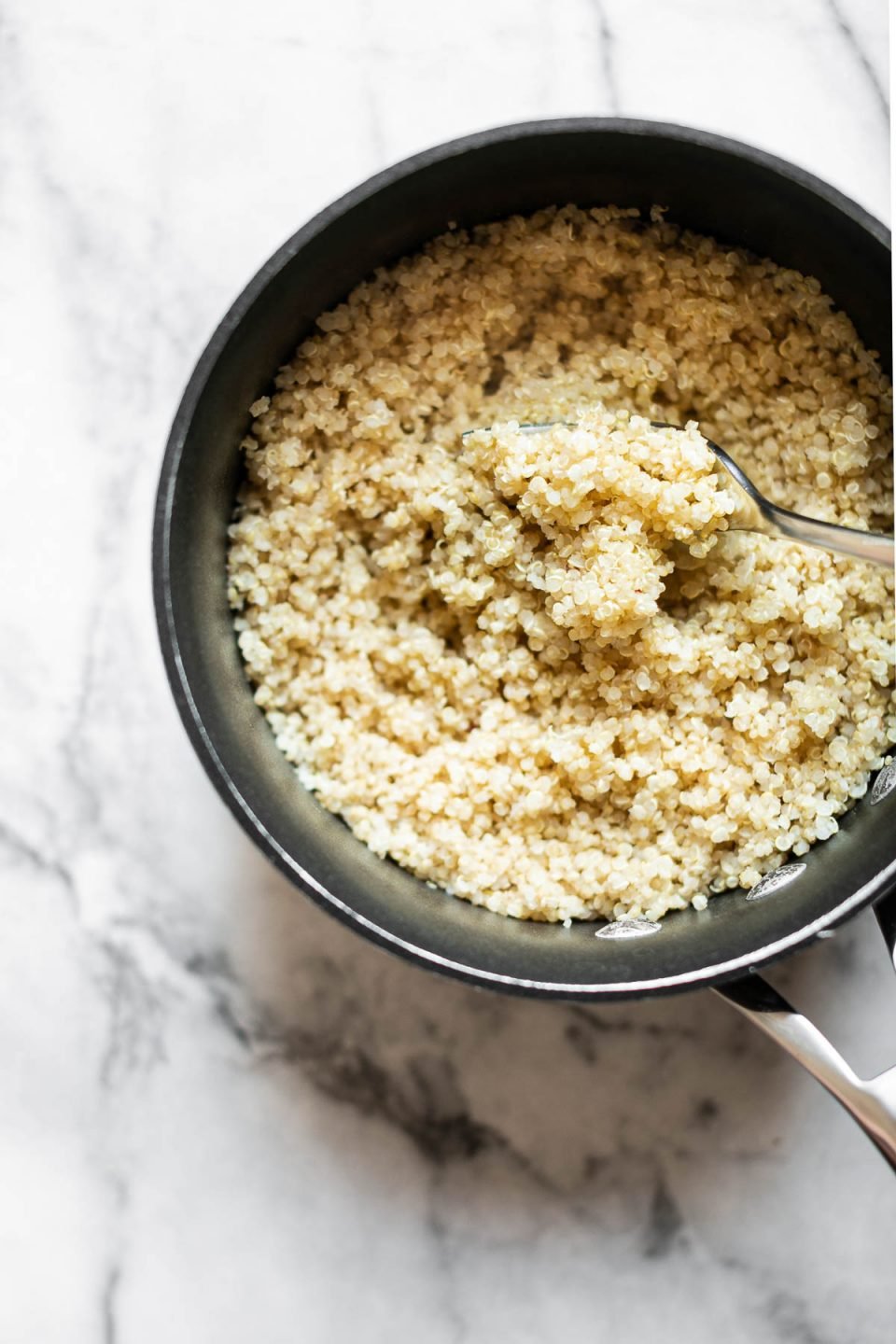 A sauce pan filled with cooked white quinoa sits on top of a white & gray marble surface. A fork that was used to fluff the perfectly cooked quinoa rests inside the sauce pan.