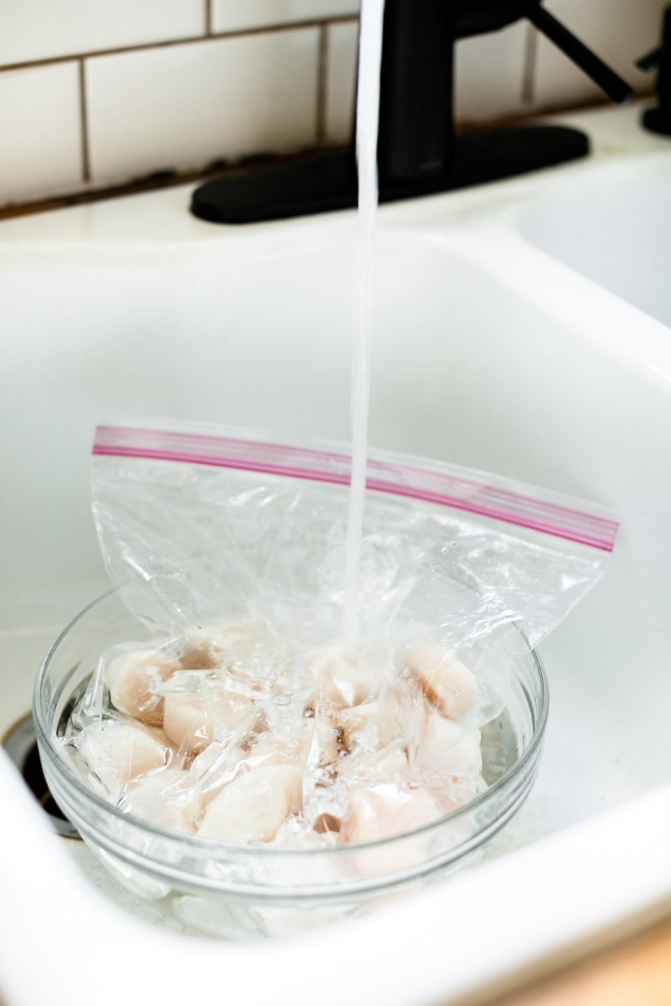How to thaw frozen scallops – Frozen scallops in a zip-top bag in a glass mixing bowl in a farmhouse-style sink with cold water streaming over them.
