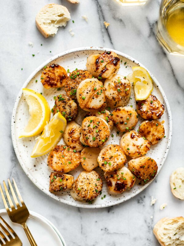 Seared scallops shown on a speckled ceramic plate, topped with freshly snipped chives & next to lemon wedges. The plate sits atop a white marbled surface surrounded by sliced, crusty bread, white wine in glasses, & gold flatware.