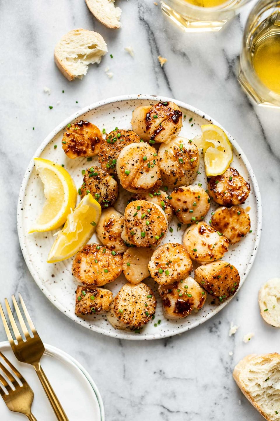 Seared scallops shown on a speckled ceramic plate, topped with freshly snipped chives & next to lemon wedges. The plate sits atop a white marbled surface surrounded by sliced, crusty bread, white wine in glasses, & gold flatware.