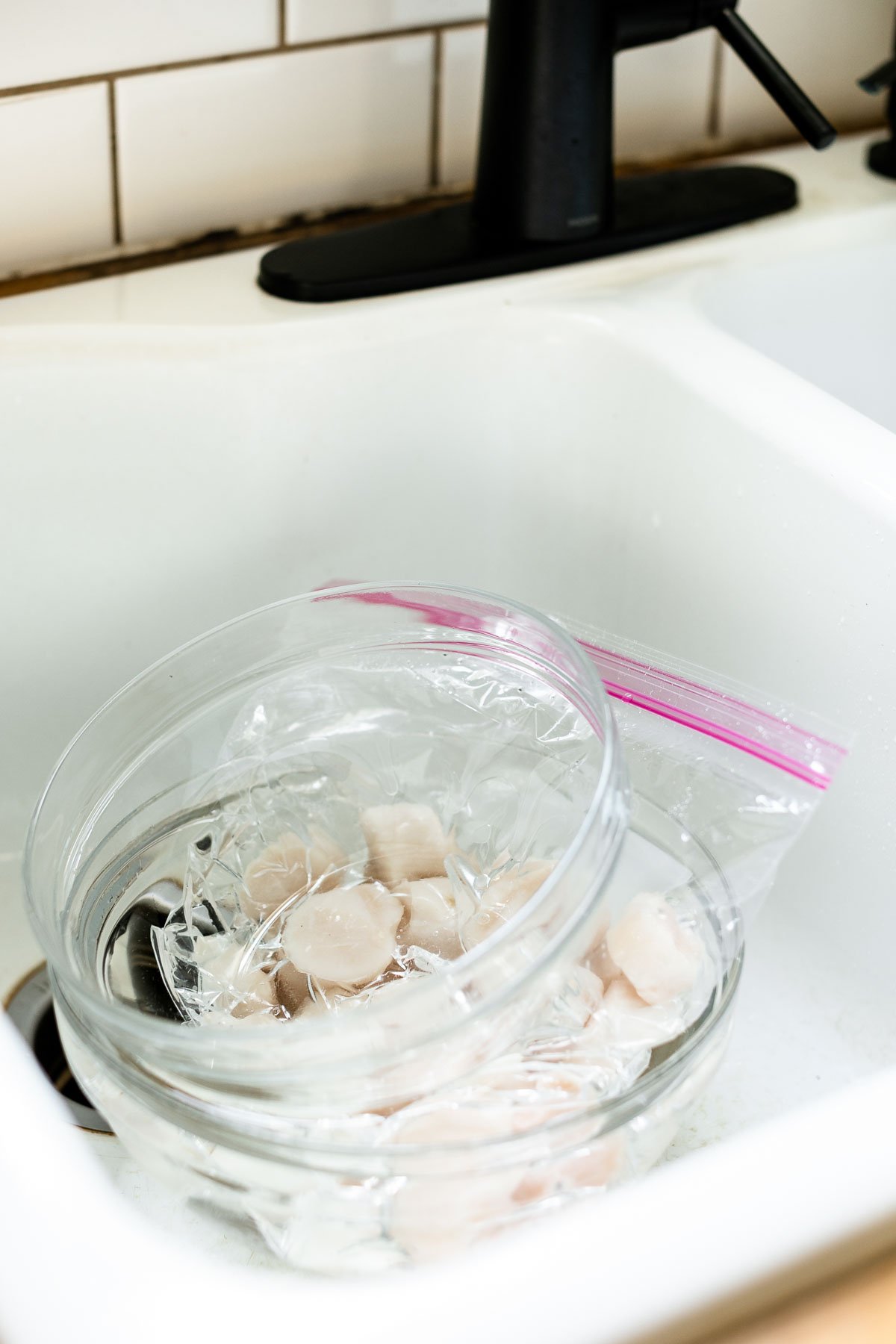 How to thaw frozen scallops – Frozen scallops in a zip-top bag in a glass mixing bowl in a farmhouse-style sink. A second glass mixing bowl sits atop the scallops, keeping them submerged in cool water.
