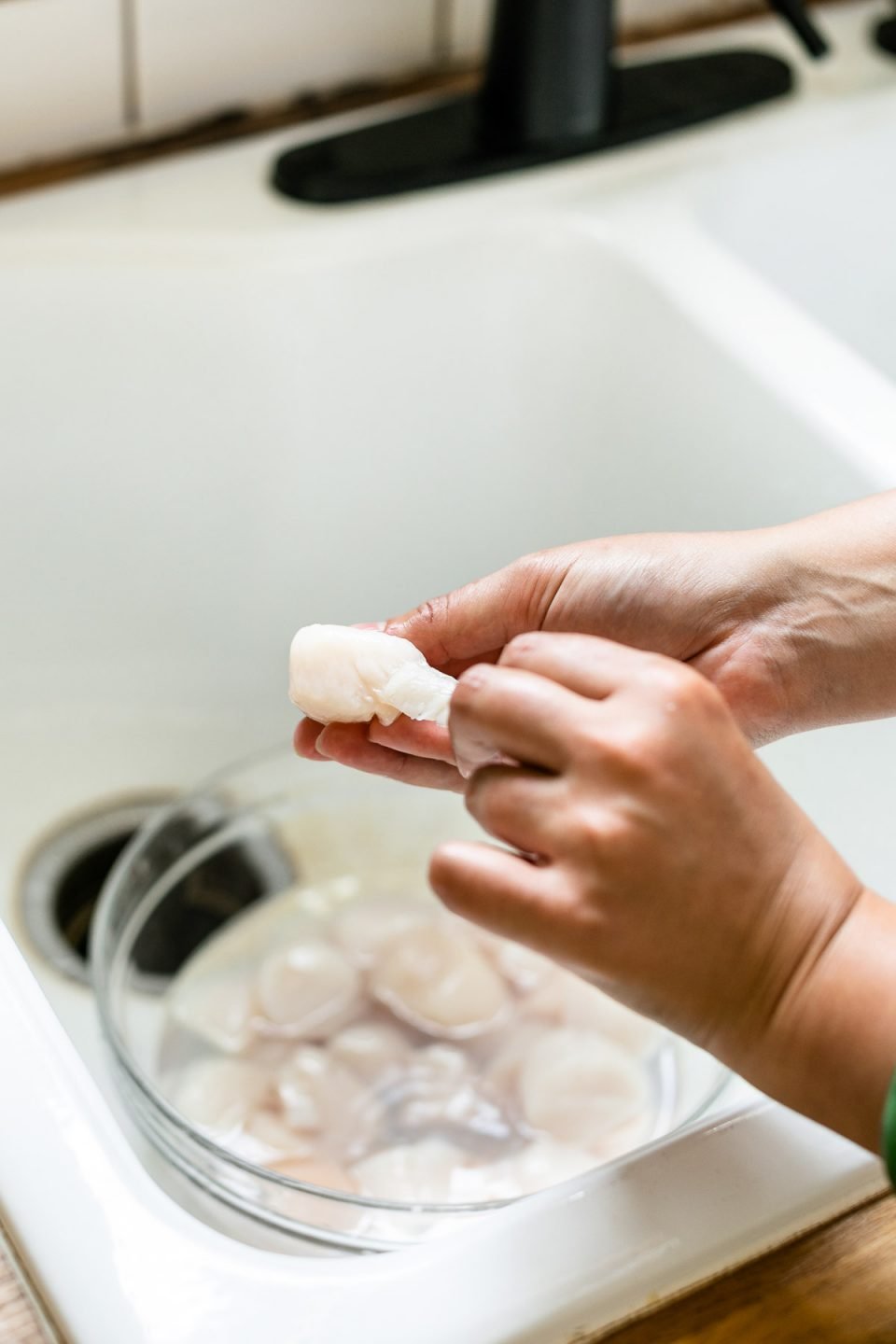 Cleaning scallops for searing – A woman's hands shown tearing the side muscle tissue away from sea scallop meat. In the background, a farmhouse-style sink with a bowl of sea scallops in it.