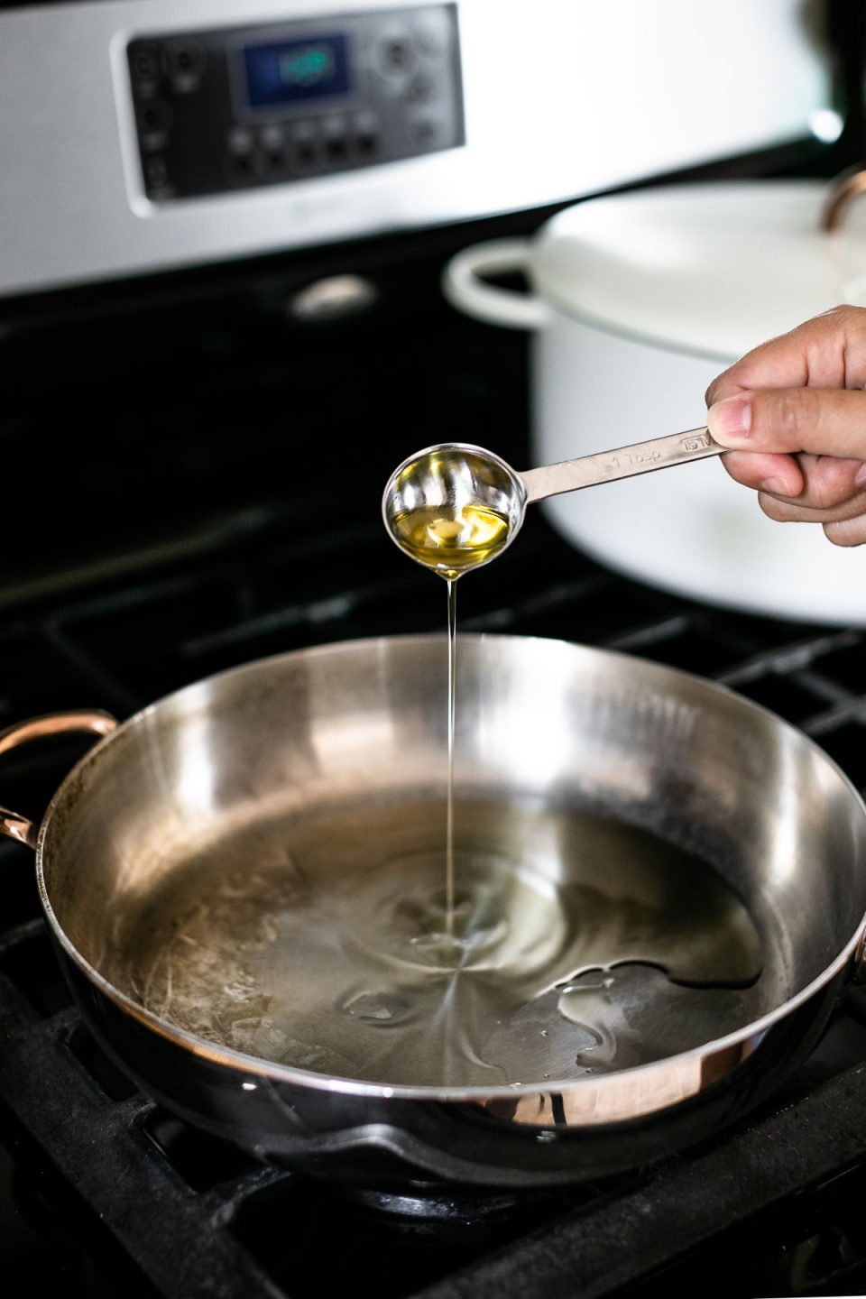 How to sear scallops, Step 1 – A woman's hand shown pouring oil into a stainless steel skillet from a tablespoon measure. The skillet sits atop a gas range with a white Dutch oven in the background.