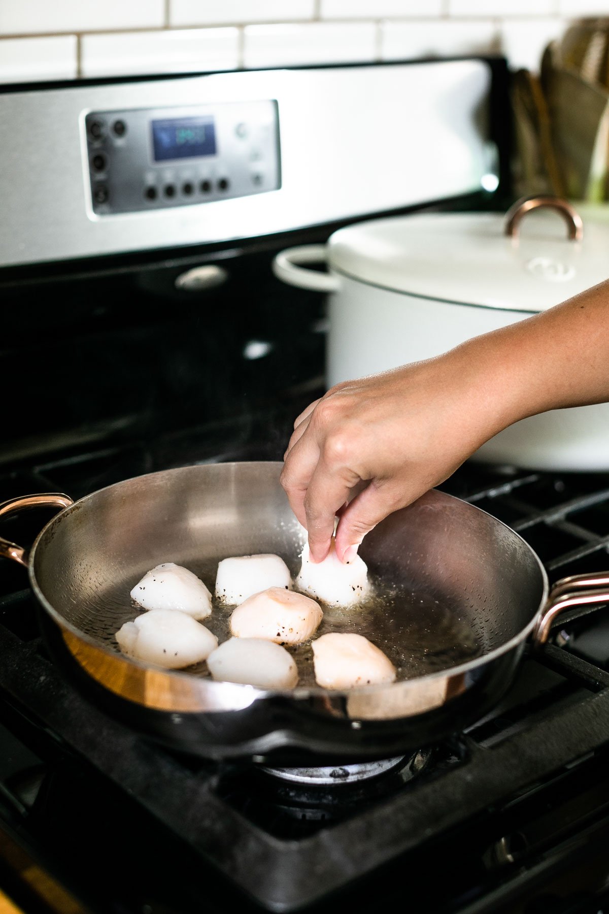 How to sear scallops, Step 3 – A woman's hand shown adding sea scallops to stainless steel skillet. The skillet sits atop a gas range with a white Dutch oven in the background.