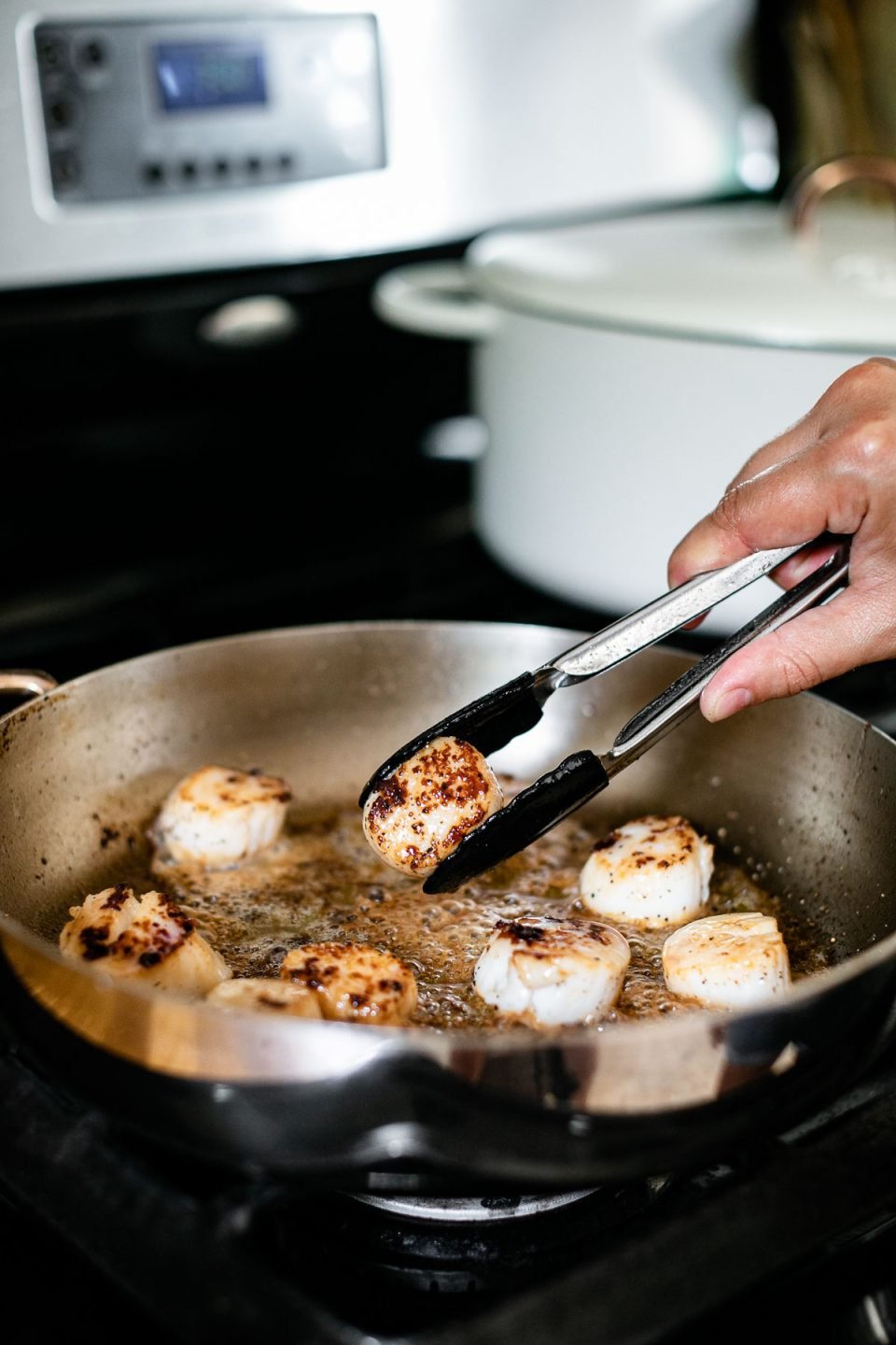 How to sear scallops, Step 3 – A woman's hand shown using small black tongs to flip seared scallops in a stainless steel skillet. The skillet sits atop a gas range with a white Dutch oven in the background.