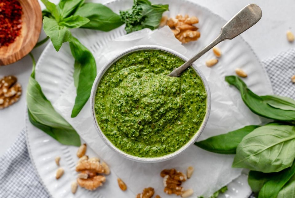 Kale pesto in a bowl atop a white plate. There is a spoon in the pesto. Surrounding the plate are walnuts, pine nuts, fresh basil & kale.