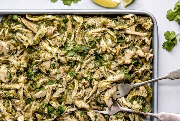 Shredded Salsa Verde Chicken (Pollo Verde) shown on a large sheet pan. 2 forks, used for shredding, are nestled to the chicken on the sheet pan. The sheet pan sits atop a light blue surface. Placed around the sheet pan are cilantro leaves & lime wedges.