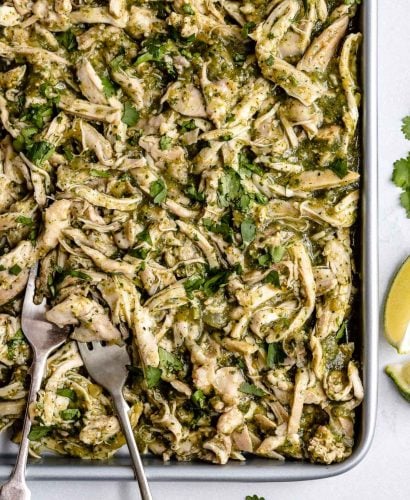 Shredded Salsa Verde Chicken (Pollo Verde) shown on a large sheet pan. 2 forks, used for shredding, are nestled to the chicken on the sheet pan. The sheet pan sits atop a light blue surface. Placed around the sheet pan are cilantro leaves & lime wedges.