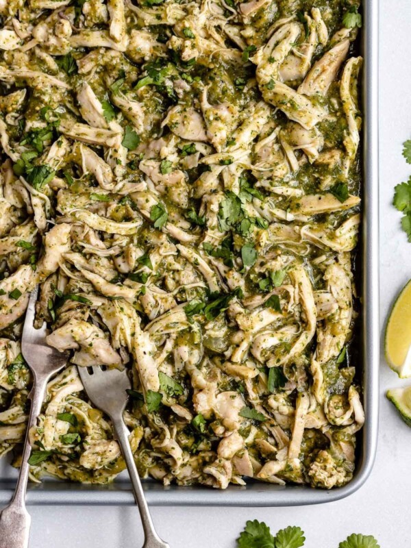 Shredded Salsa Verde Chicken (Pollo Verde) shown on a large sheet pan. 2 forks, used for shredding, are nestled to the chicken on the sheet pan. The sheet pan sits atop a light blue surface. Placed around the sheet pan are cilantro leaves & lime wedges.