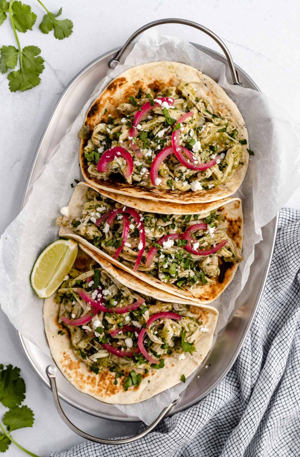 Shredded salsa verde chicken (pollo verde) shown in 3 tacos. The tacos are topped with chopped cilantro and pickled red onions, placed on a metal tray with handles. The tray sits atop a light blue surface, next to a checkered blue linen napkin & a few cilantro leaves.