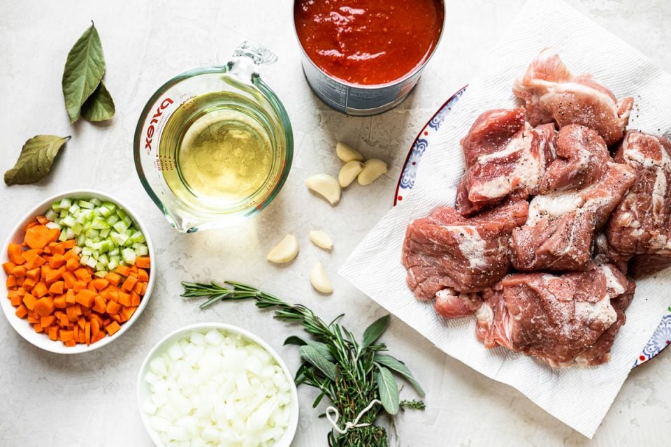 Pork ragu ingredients arranged on a white surface: 6 large pieces of pork shoulder, onions, carrots, celery, bay leaves, garlic, fresh herbs, wine & crushed tomatoes.