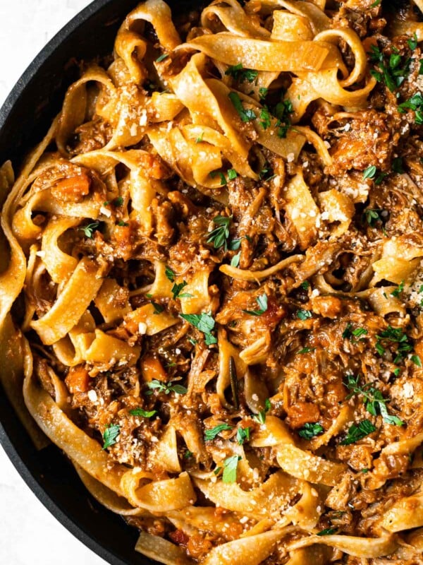 Close up of pork shoulder ragu tossed into pappardelle pasta in a skillet. The pasta is topped with fresh herbs.