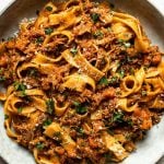 Pork ragu pappardelle shown in a large gray pasta bowl. The pasta is topped with fresh herbs. Surrounding the bowl is a small bowl of grated parmesan & some fresh thyme sprigs.