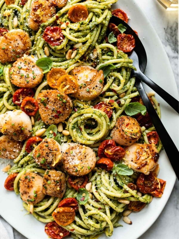 A close up of Seared Scallops and Kale Pesto pasta on a white serving platter. Black serving ware rests on the platter & is tucked into the pasta ready to serve. The platter sits on a grey and white marble surface. A light grey linen napkin is tucked under the platter in the foreground and a clear glass of white wine is partially shown in the background.