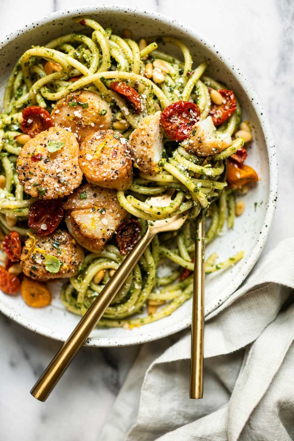 Seared Scallops and Kale Pesto pasta in a black & white speckled pasta bowl. A gold fork & spoon rests inside the bowl & is tucked into the pasta ready to serve. The bowl sits on a grey and white marble surface. A light grey linen napkin is tucked under the platter in the foreground.