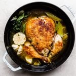 Browned whole chicken in a large white Dutch oven with soup aromatics before roasting. The pot is filled with chicken broth.