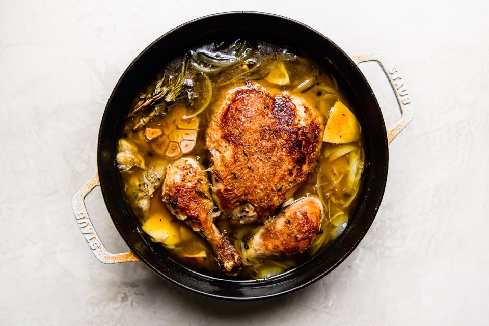 Whole roasted chicken soup after roasting. A whole roasted chicken sits in a large white Dutch oven with a rich yellow broth & spent herbs, lemon wedges & garlic.