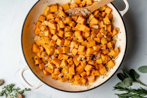 Sauteeing butternut squash in a large skillet with onion, sage, & garlic. A wooden spoon is nestled into the squash. The skillet sits atop a light blue surface, next to fresh herbs & whole nutmeg.