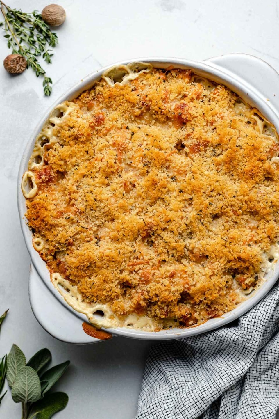 Baked butternut squash au gratin in a round baking dish. The baking dish sits atop a light blue surface, next to a checkered linen napkin, fresh herbs & whole nutmeg.