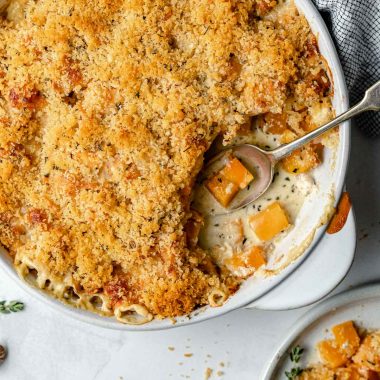 Baked butternut squash au gratin in a round baking dish. A spoonful of gratin has been taken out of the baking dish. The baking dish sits atop a light blue surface, next to a checkered linen napkin, fresh herbs & whole nutmeg.