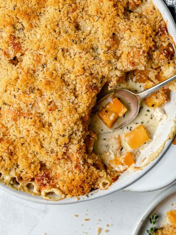 Baked butternut squash au gratin in a round baking dish. A spoonful of gratin has been taken out of the baking dish. The baking dish sits atop a light blue surface, next to a checkered linen napkin, fresh herbs & whole nutmeg.